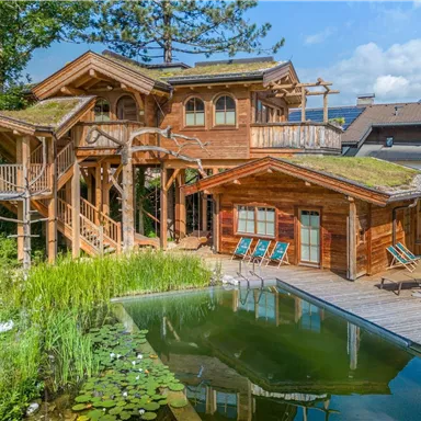 Ein schönes, modernes Holzhaus mit Balkon und großem Garten. Im Vordergrund befindet sich ein ruhiger Teich umgeben von Pflanzen und Liegestühlen.