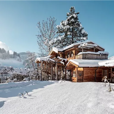 Ein malerisches Holzhaus im Schnee, umgeben von verschneiten Bäumen. Der klare blaue Himmel bildet einen schönen Kontrast zu der winterlichen Landschaft.