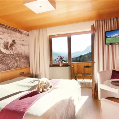 Ein helles Hotelzimmer mit einem Doppelbett und Holzwänden. Durch das Fenster ist eine Berglandschaft zu sehen.