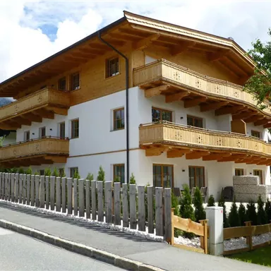 A modern, multi-story building with wooden cladding and balconies. Surrounded by green trees and a well-kept garden fence.
