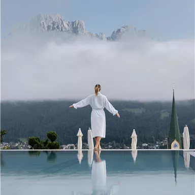 A woman in a bathrobe stands at the edge of an infinity pool. In the background, mountains and fog are visible, creating a calm and relaxing atmosphere.