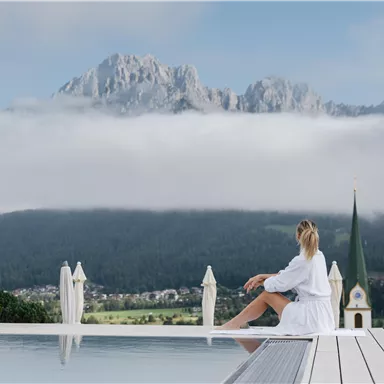 A woman sits at the edge of an infinity pool and looks at majestic mountains surrounded by clouds. In the background, there is a picturesque landscape with a church tower visible.