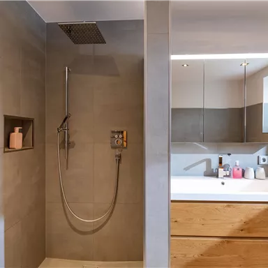 Ein modernes Badezimmer mit einer Dusche und einer großen Badewanne aus Holz. Die Wände sind in neutralen Grautönen gestaltet und es gibt eine große Fensterfront für natürliches Licht.