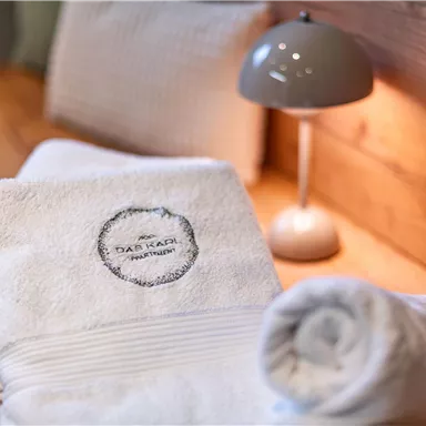 Ein weicher, weißer Handtuchstapel mit einem Logo. Im Hintergrund steht eine Tischlampe auf einem Holzregal.