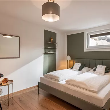 Ein modernes Schlafzimmer mit einem Doppelbett und minimalistischer Einrichtung. Das Licht ist hell und freundlich, mit einem gemütlichen Beistelltisch und einer Lampe.