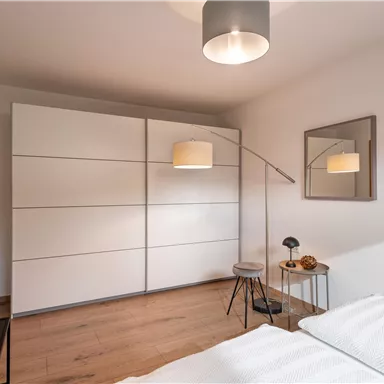Ein modernes Schlafzimmer mit einem großen weißen Kleiderschrank und einem hölzernen Boden. Es gibt eine Stehlampe und einen Spiegel an der Wand.
