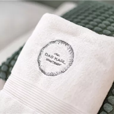 Ein weißes Handtuch mit dem Logo von "Das Kaiser Wasser" liegt auf einem grünen Kissen. Der Hintergrund ist sanft und hell gehalten.