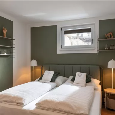 Ein modernes Schlafzimmer mit zwei Betten und minimalistischem Design. Die Wände sind in sanften Grüntönen gehalten und es gibt Nachttische mit Leuchten.