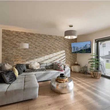 Ein modernes Wohnzimmer mit einer bequemen Couch und einem runden Tisch. Große Fenster lassen viel Licht herein und ein Fernseher zeigt eine Naturaufnahme.