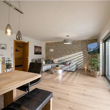 Ein modernes Wohnzimmer mit einer großen Fensterfront und einem Essbereich. Die stilvolle Einrichtung umfasst eine gemütliche Couch und dekorative Pflanzen.