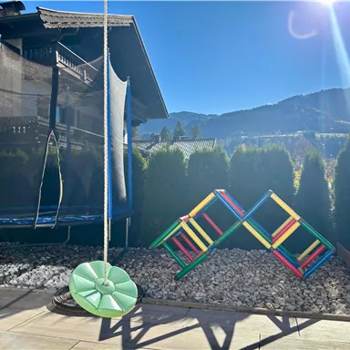 A garden with a trampoline and a swing seat. In the background, green trees and mountains can be seen.