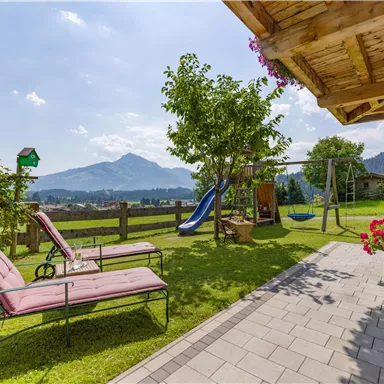 An inviting garden with loungers and a playground. In the background, mountains rise under a clear sky.