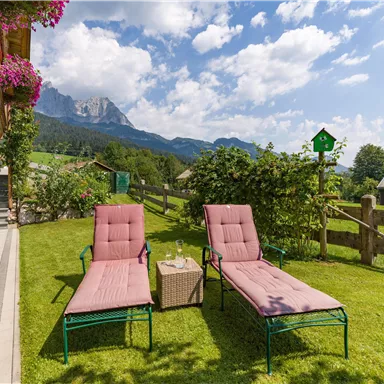A relaxed garden landscape with two loungers and a small table. In the background, green meadows and mountains can be seen under a blue sky.