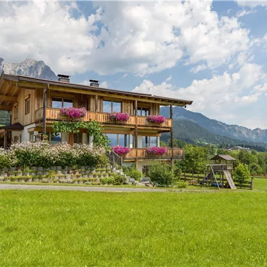 A beautiful wooden house with flowered balconies in a green meadow landscape. In the background, there are mountains and a blue sky.