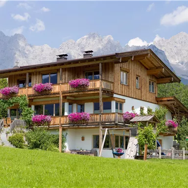 A beautiful wooden house with blooming balconies and a green lawn in front. In the background, majestic mountains can be seen.