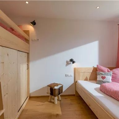 A cozy bedroom with a loft bed and a single bed. It features warm wooden furnishings and red cushions with a cheerful pattern.
