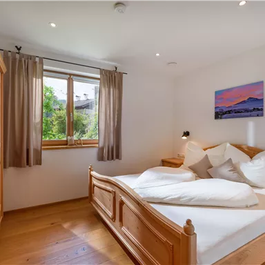 A cozy bedroom with a large bed and wooden furniture. Daylight falls through the window and looks out onto a green landscape.