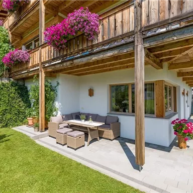 A cozy outdoor area with a table and chairs under a wooden overhang. The area is surrounded by green grass and blooming plants.