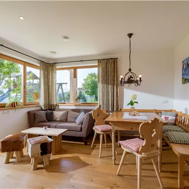 A cozy living area with large windows and wooden furniture. The room is bright and inviting, with a seating area and a dining table.