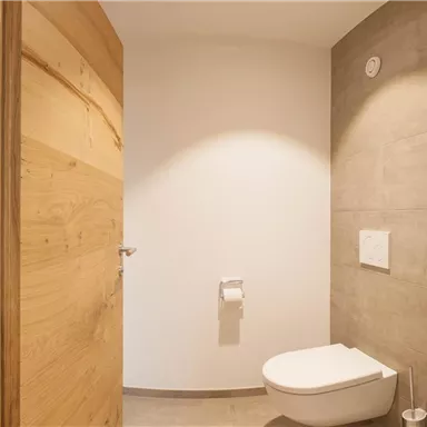 Ein modernes Badezimmer mit einer weißen Toilette und hellen Wänden. Die Tür aus Holz verleiht dem Raum eine warme Atmosphäre.