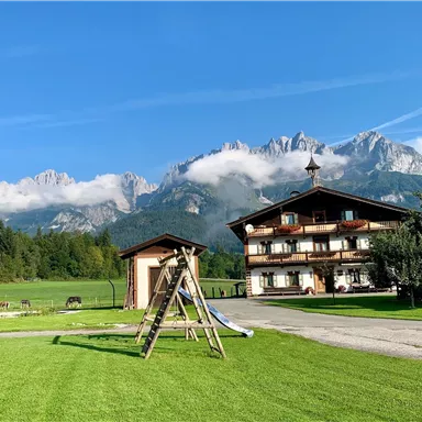 Ein malerisches Bergpanorama mit hohen Gipfeln und etwas Nebel. Im Vordergrund steht ein Spielplatz und ein charmantes Gebäude mit Balkonen.