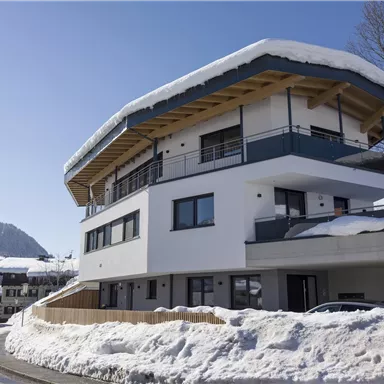 A modern house with a snow-covered roof in a winter landscape. The surroundings show a quiet road and snow-covered mountains in the background.