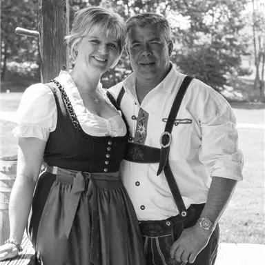 Ein Paar in traditioneller Tracht steht lächelnd vor einer ländlichen Kulisse. Der Mann trägt eine Lederhose und die Frau ein Dirndl.