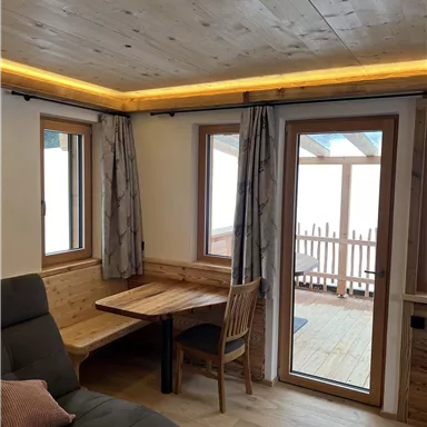 Ein gemütlicher Raum mit Holzdecke und großen Fenstern. Ein Tisch mit Stuhl steht neben einem bequemen Sofa.