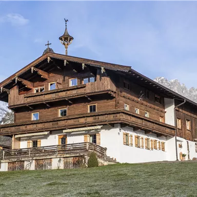 Ein traditionelles Holzhaus in den Bergen mit einem klaren Himmel im Hintergrund. Umgeben von grünen Wiesen und schneebedeckten Gipfeln.