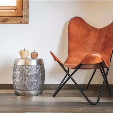 A stylish chair made of brown leather stands in a bright room next to a decorative table. On the table, there are two small fruit decorations.