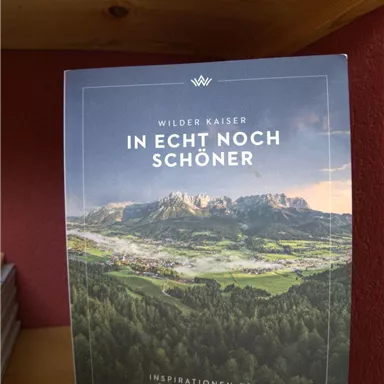 A book titled "Even More Beautiful in Reality" is on a shelf. It showcases a breathtaking landscape of the Wilder Kaiser.