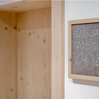 Ein Innenraum mit einem Holzrahmen, der ein Schild mit der Aufschrift „Deluxe Alpin 214“ zeigt. Die Wand ist hell und die Holzelemente verleihen dem Raum eine gemütliche Atmosphäre.