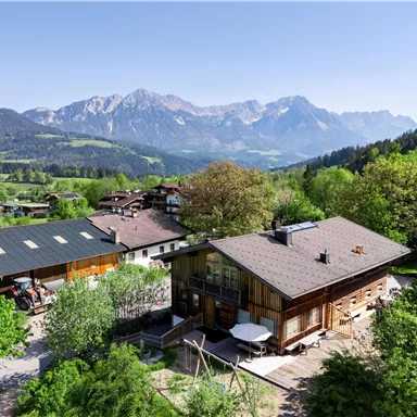 A picturesque mountain village with traditional wooden houses and lush greenery. In the background, impressive mountains rise under a clear blue sky.
