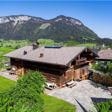 A traditional wooden house with a beautiful terrace is surrounded by green meadows. In the background, majestic mountains rise under a clear blue sky.