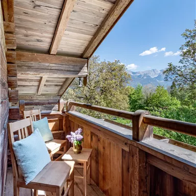 A cozy balcony with two wooden stools and a vase with flowers. In the background, trees and mountains can be seen under a clear blue sky.