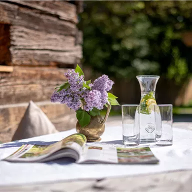 A table outdoors with a vase of flowers and a jug of water. On it also lie a magazine and some glasses.