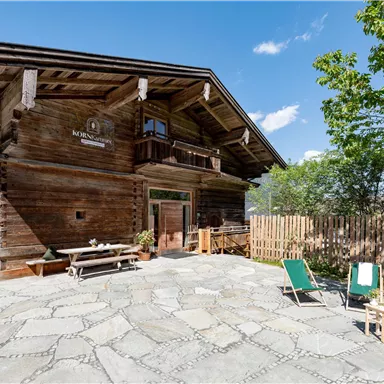 A charming wooden house with a large paved outdoor area. There are sun loungers and green foliage in the surroundings.