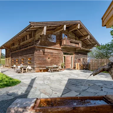 A traditional wooden house in a picturesque landscape. The terrace is furnished with wooden chairs and tables, surrounded by green meadows and trees.