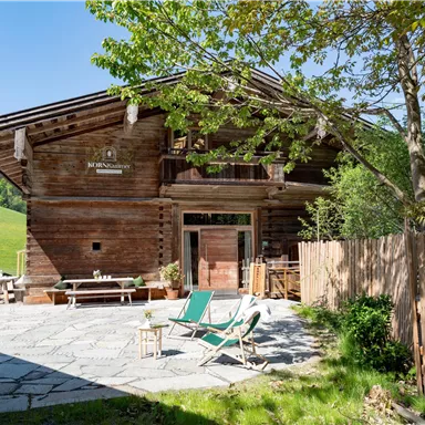 A charming wooden-clad house in a green landscape. In front of the house are lounge chairs and a paved area.