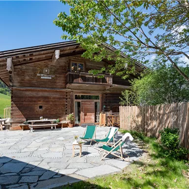 A cozy wooden house surrounded by green nature. In front of the house, there are sun loungers on a paved area.