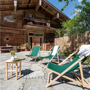 A cozy terrace with lounge chairs in front of a wooden house. Surrounded by greenery and a clear blue sky.