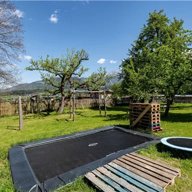 A garden with a large trampoline and a smaller trampoline nearby. The surroundings are green with trees and a hilly landscape in the background.