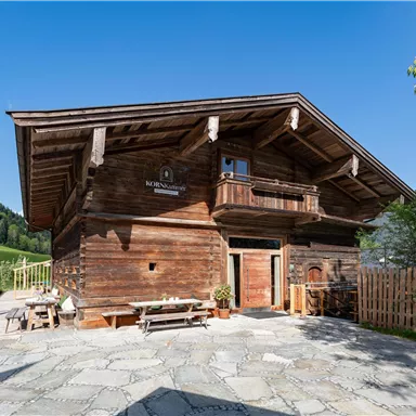 A rustic wooden house with a large balcony, surrounded by lush greenery. The clear sky and well-maintained outdoor area give the place a cozy atmosphere.