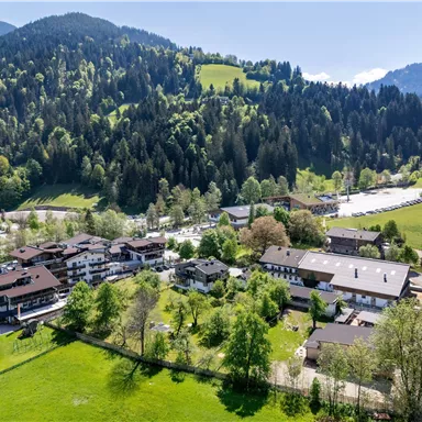 A picturesque landscape with green meadows and wooded hills. In the foreground, several traditional buildings can be seen that belong to a rural community.