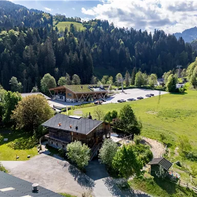 A picturesque landscape with a rustic wooden building and plenty of greenery. In the background, wooded hills rise under a blue sky.