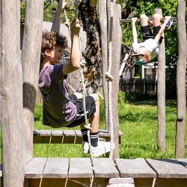 Two children are playing in a playground. One is swinging on a swing, while the other is hanging upside down from a pole.