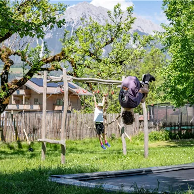 A playground with two children playing on a swing and a horizontal bar. In the background, trees and mountains are visible.