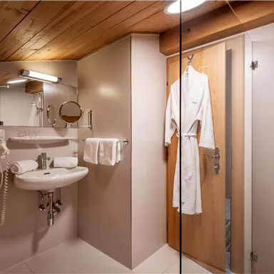 A modern bathroom with a shower cubicle, a sink, and a wooden shelf. A bathrobe hangs on the door, and the room is comfortably furnished.