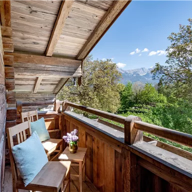 A cozy balcony with wooden cladding and two chairs. In the background, green trees and the mountains can be seen.