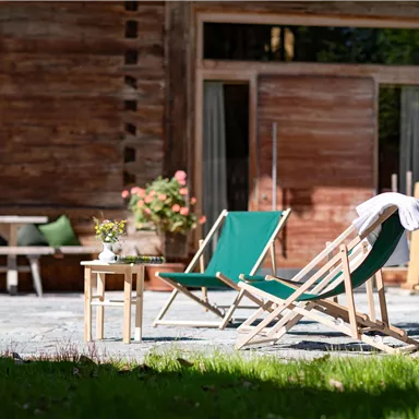 A relaxed outdoor area with two lounge chairs and a small table. In the background, a wooden house and green plants can be seen.